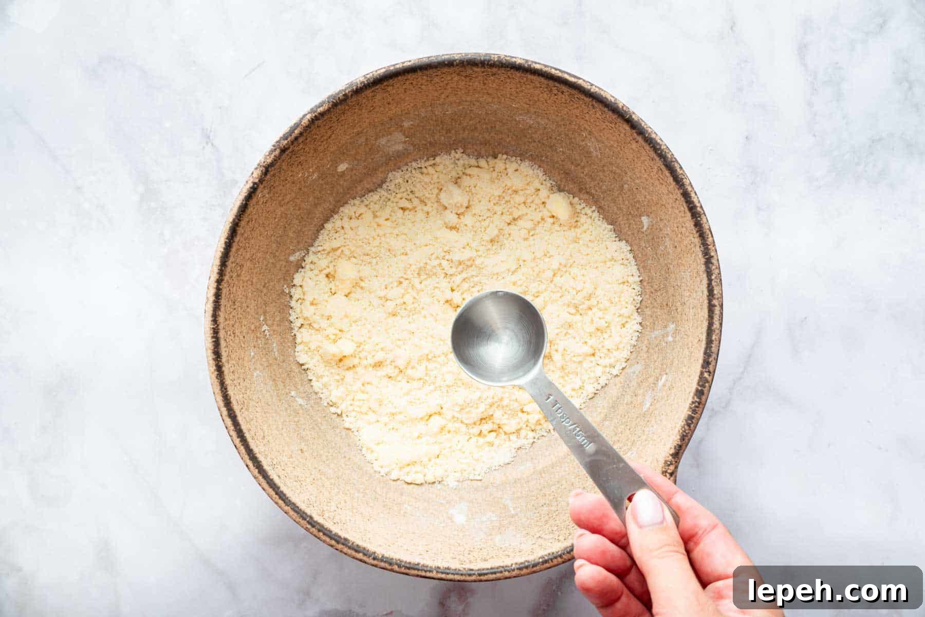 Hand adding one teaspoon of clear liquid to bowl of crumbly pastry.