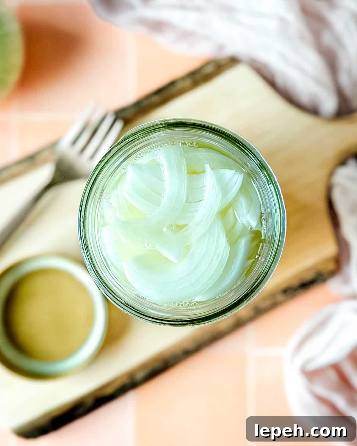 Pickled white onions in a glass jar on a wooden cutting board surrounded by white onions, a fork, and a pink linen.