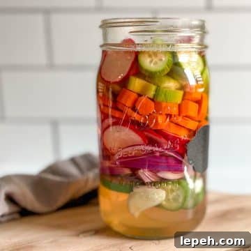 A glass jar of mixed pickles sits on a wooden cutting board.