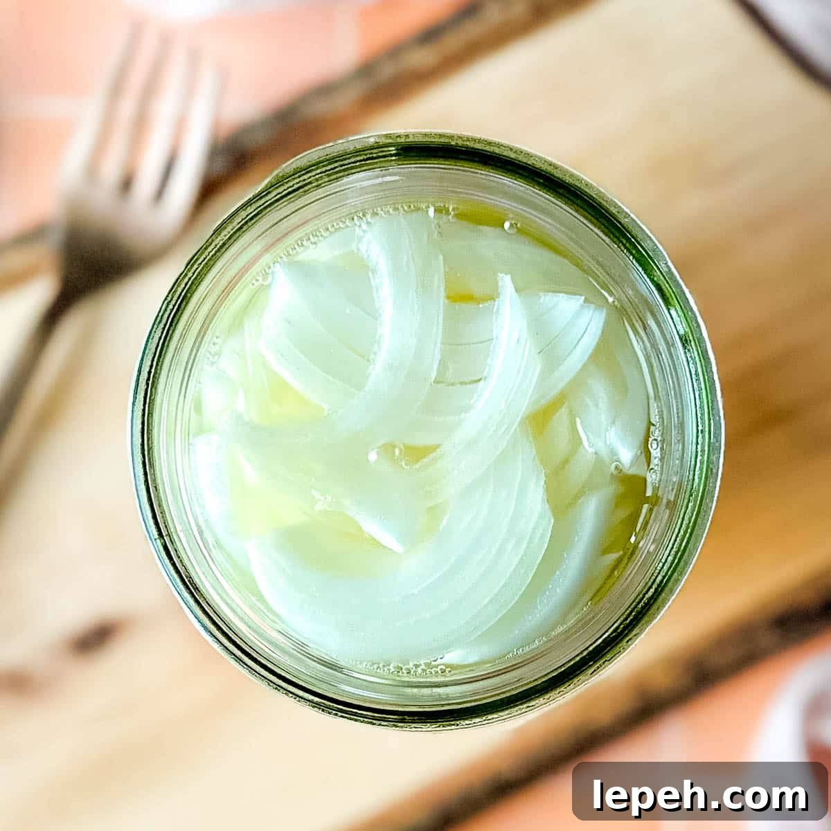 Pickled white onions in a glass jar on a wooden cutting board surrounded by white onions, a fork, and a pink linen.