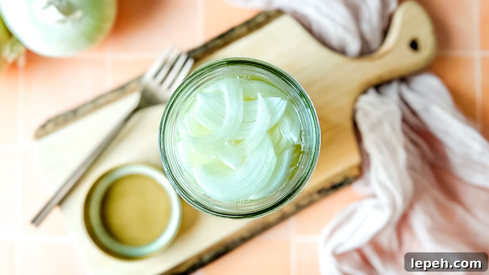 Pickled white onions in a glass jar on a wooden cutting board surrounded by white onions, a fork, and a pink linen.