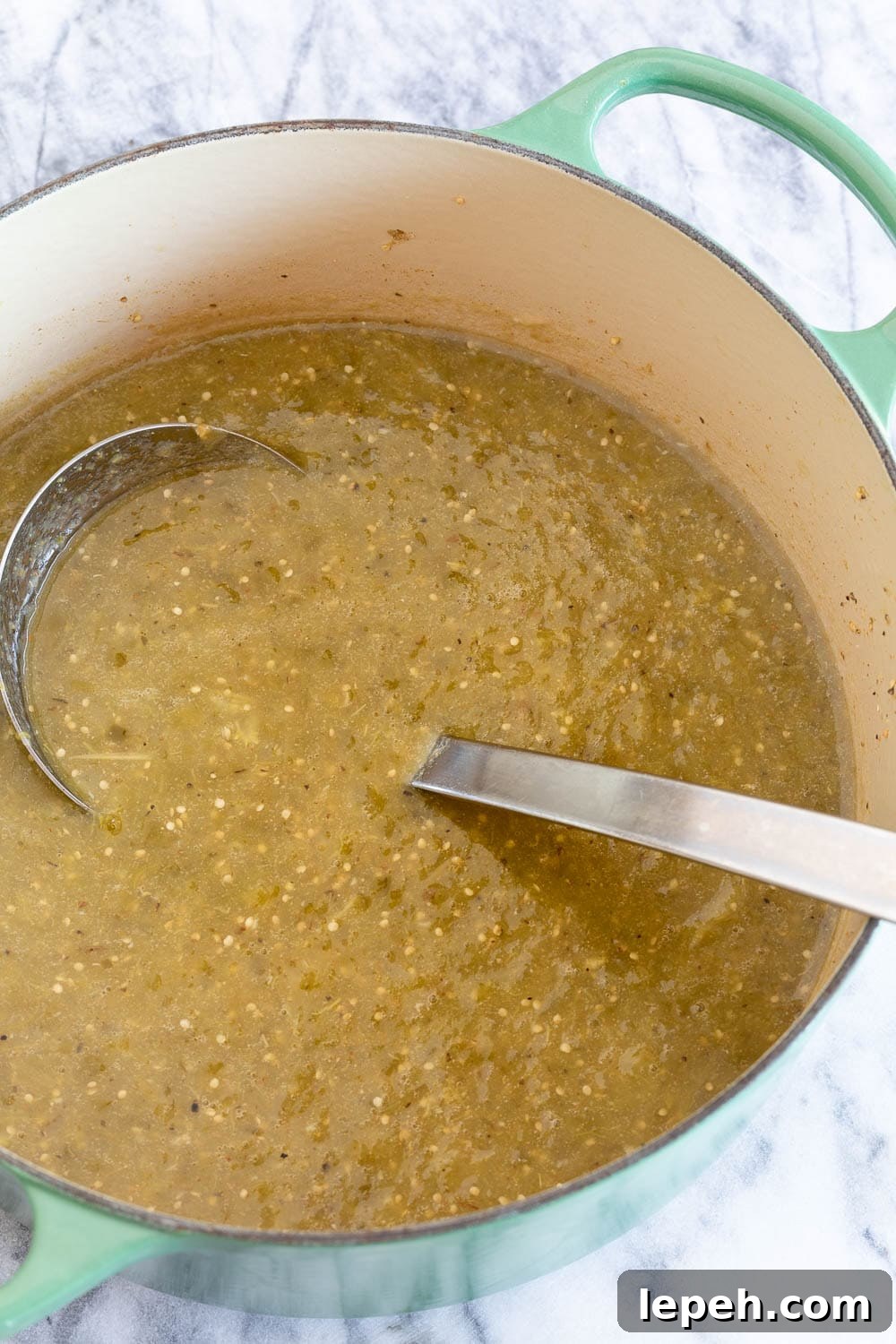 A pot of finished green tomatillo sauce with a ladle ona marble counter.