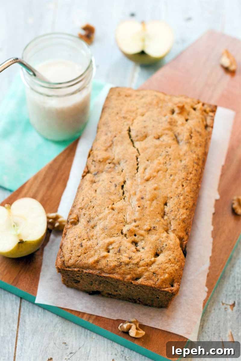 Top-down vertical shot of a loaf-shaped apple quick bread on a piece of parchment paper on top of a rectangular serving board, with sliced fruit, a jar of almond milk with a stainless steel straw, on a blue cloth background.