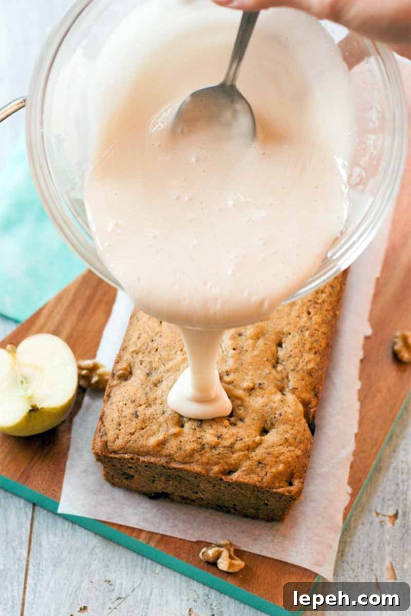 Vertical image of a hand holding a glass bowl of sugar glaze with a spoon in it, and pouring it onto a breakfast apple cake, on top of a piece of parchment on a brown wood platter, with half of an apple, on a folded light blue cloth on top of a white painted wood surface.