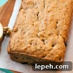Closeup closely cropped image of a loaf-shaped breakfast cake, on a rectangular piece of parchment on a slightly larger brown wood board, on a pastel aqua-colored cloth on top of a white wood surface, with half an an apple to the left that is cut side up.