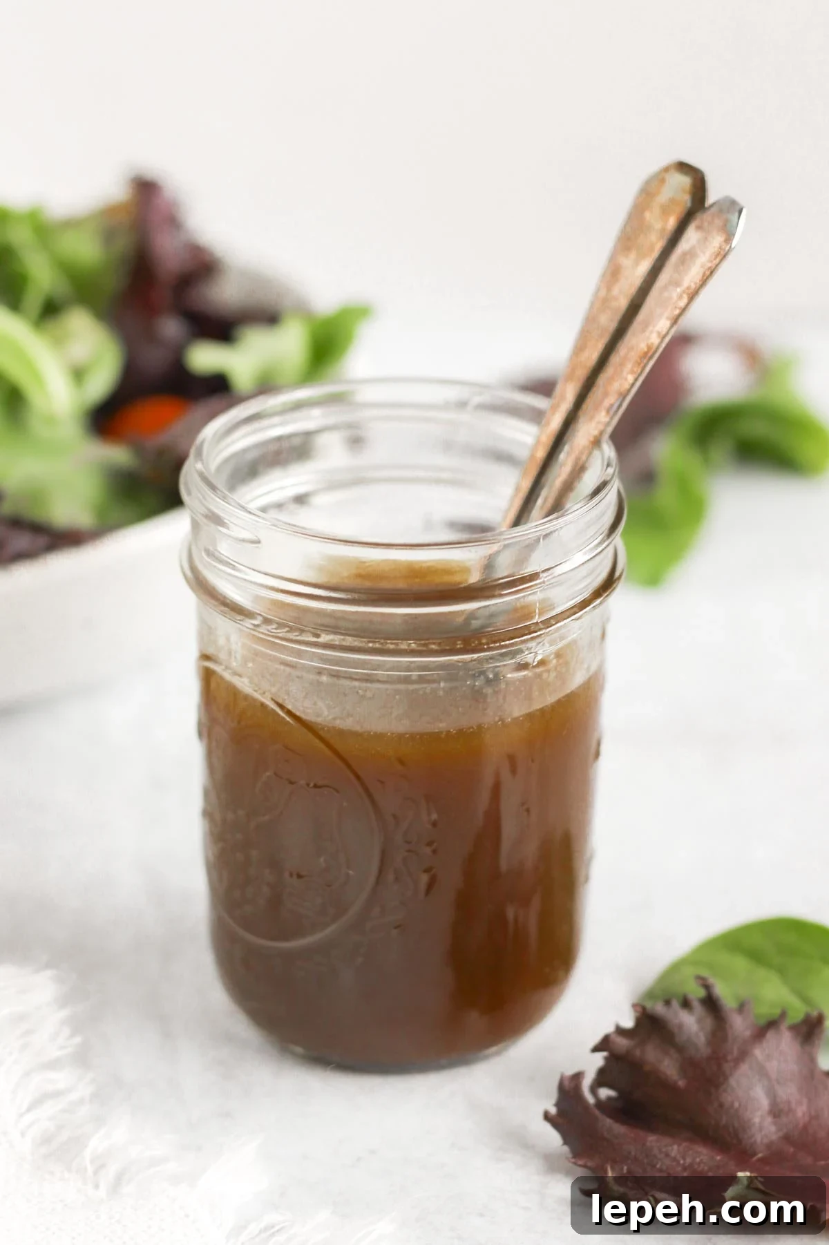 Mason jar with two spoons filled with balsamic vinaigrette with a bowl of mixed greens in the background.