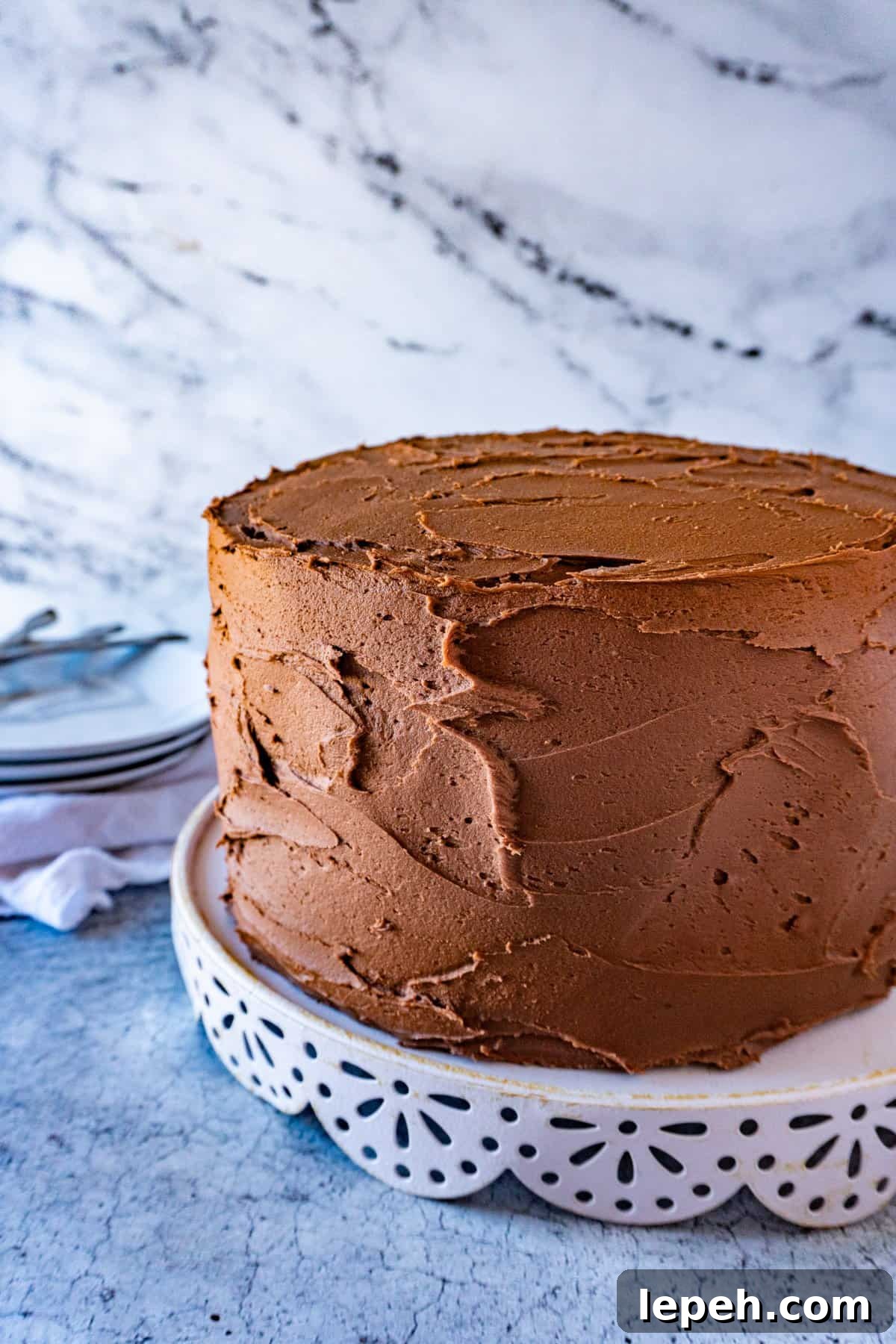  Buttermilk chocolate cake on white cake stand with plates and forks in background.