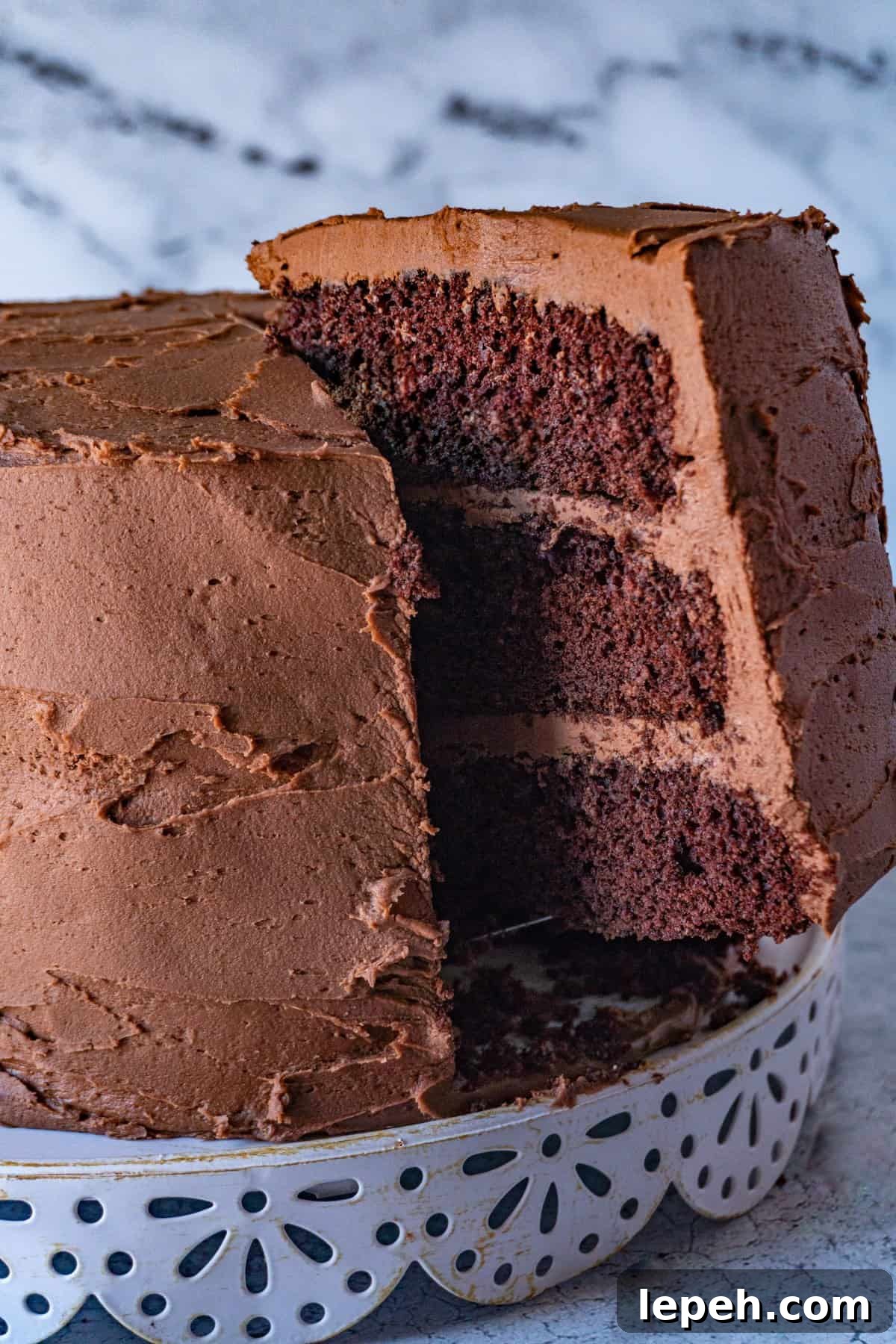 lifting a slice of buttermilk chocolate cake from the entire cake, sitting on a white cake stand.