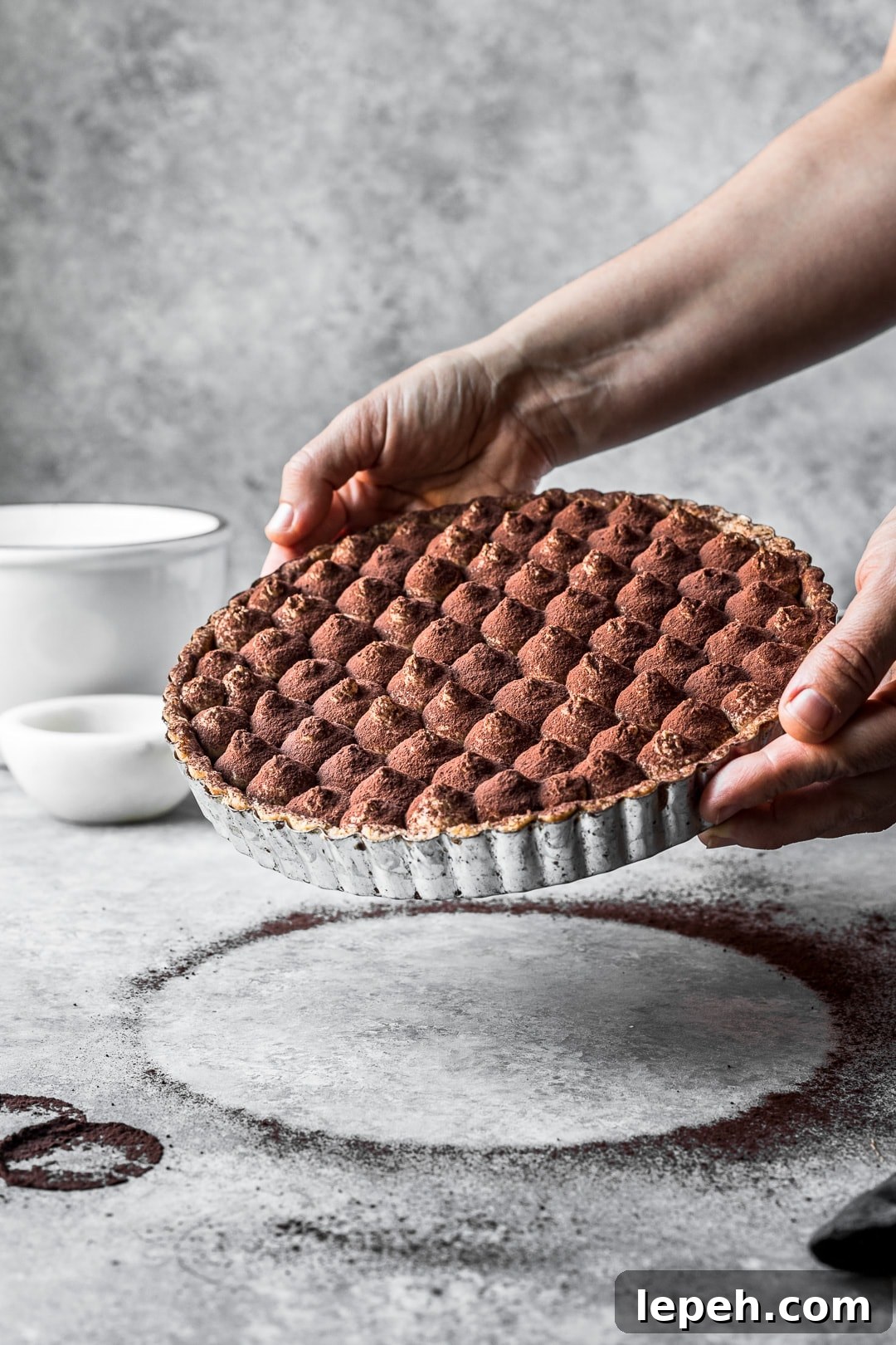 Hands holding a chocolate coffee mascarpone tart for display against a grey background