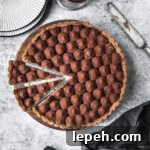 Two slices are cut out of a chocolate tart. It rests on a round wooden cutting board on a grey stone surface. There are plates, a knife and a napkin nearby.
