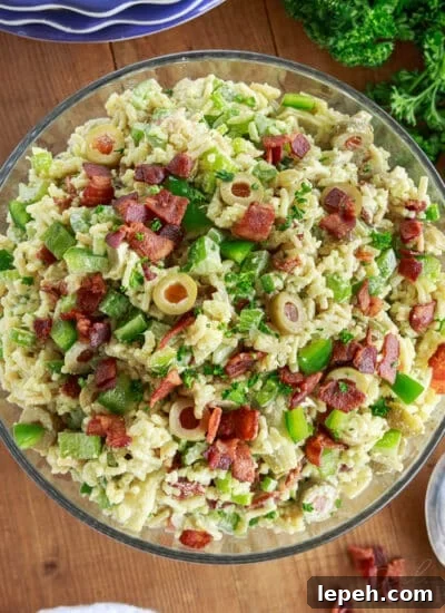 Prepared bowl of Rice a roni salad on a wooden board with stack of plates, bunch of parsley and green onions, and a spoon and linen.