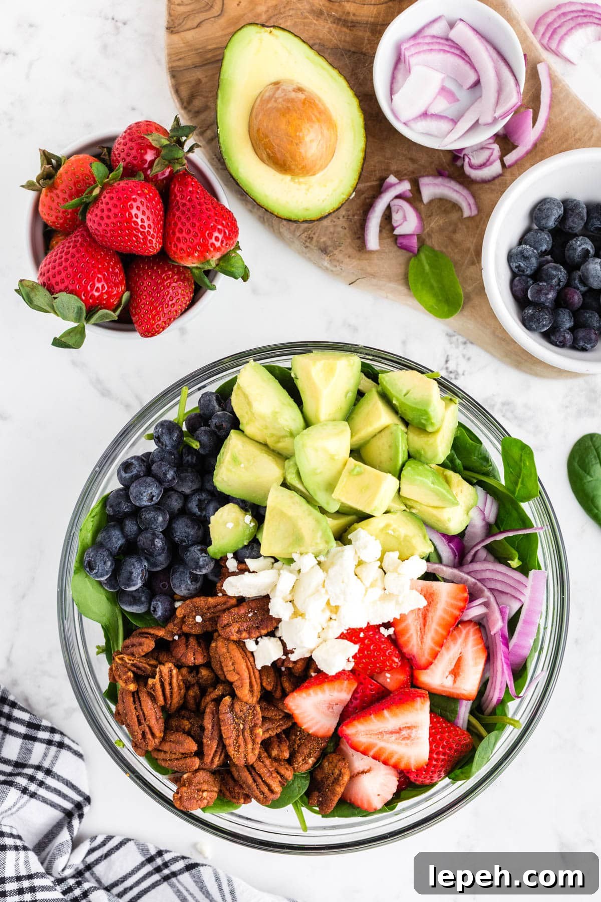 Glass bowl filled with spinach, pecans, cut strawberries, cut avocado, blueberries, onions, feta cheese, bowl with strawberries, half an avocado, bowl with blueberries, bowl with onions, black and white checked linen on a white marble countertop.