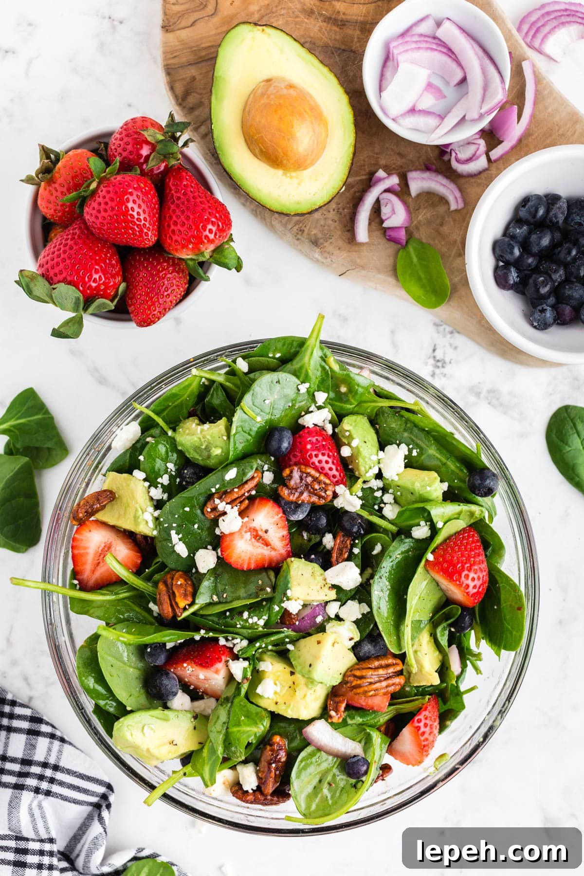 Glass bowl filled with tossed Strawberry Feta Spinach Salad, bowl with strawberries, half an avocado, bowl with blueberries, bowl with onions, black and white checked linen on a marble countertop.