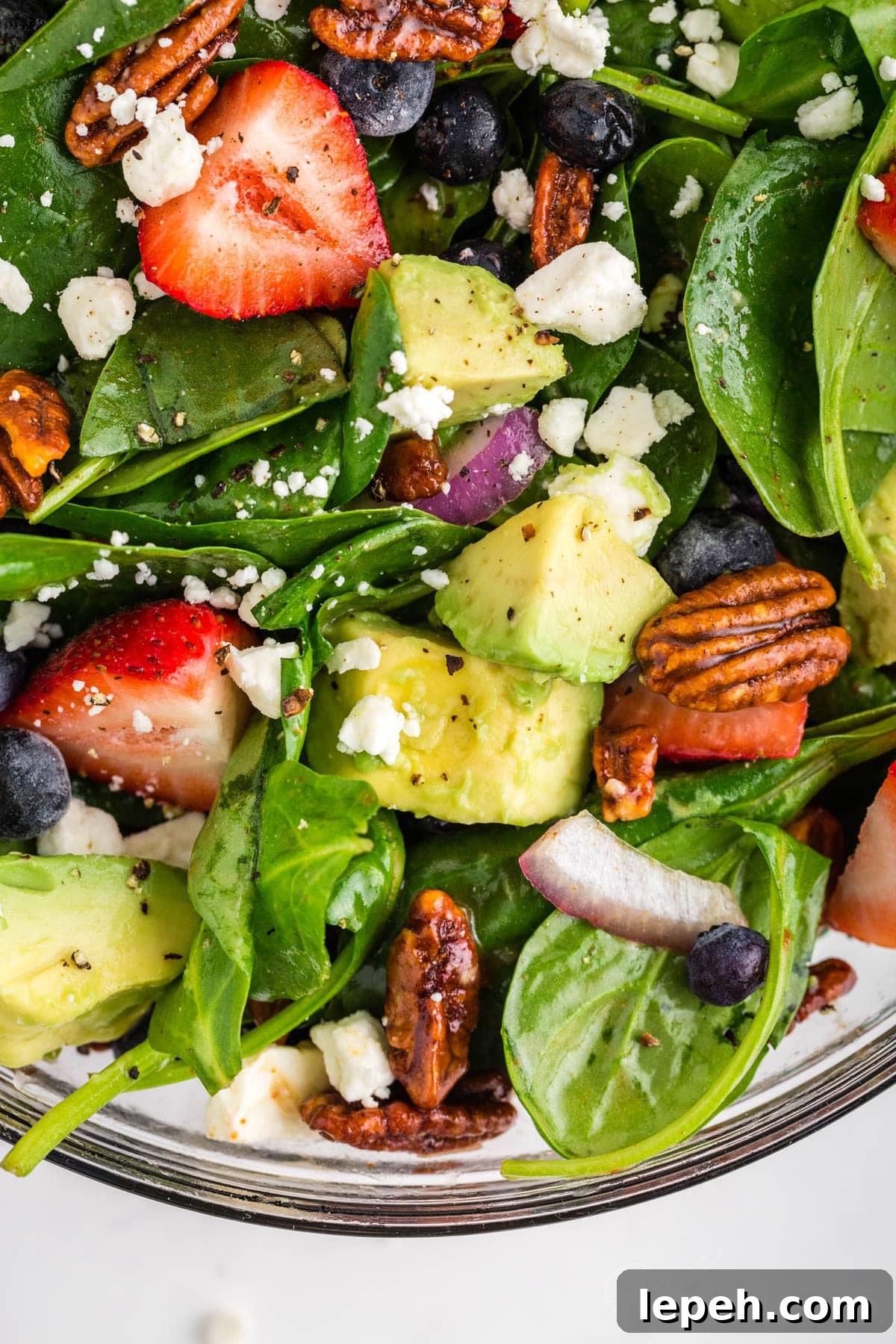 Chunks of avocado, sliced strawberries, feta cheese crumbles, red onion pieces and candied pecans over a bed of spinach in glass bowl.