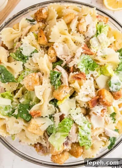 Large glass serving bowl filled with chicken caesar pasta salad with wooden salad spoons and fresh lemons in the background, all placed on a white marble surface.
