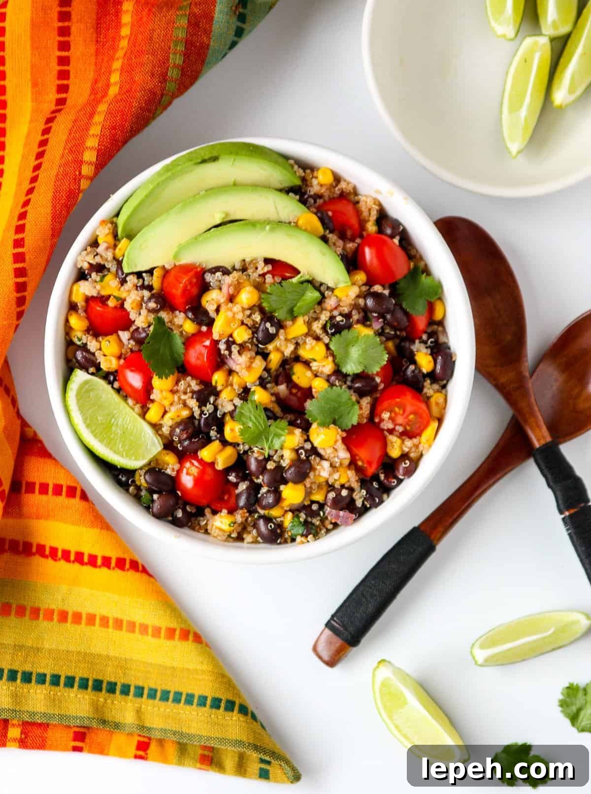 A bowl of salad on a white table with two wooden salad spoons and a small dish of lime wedges.
