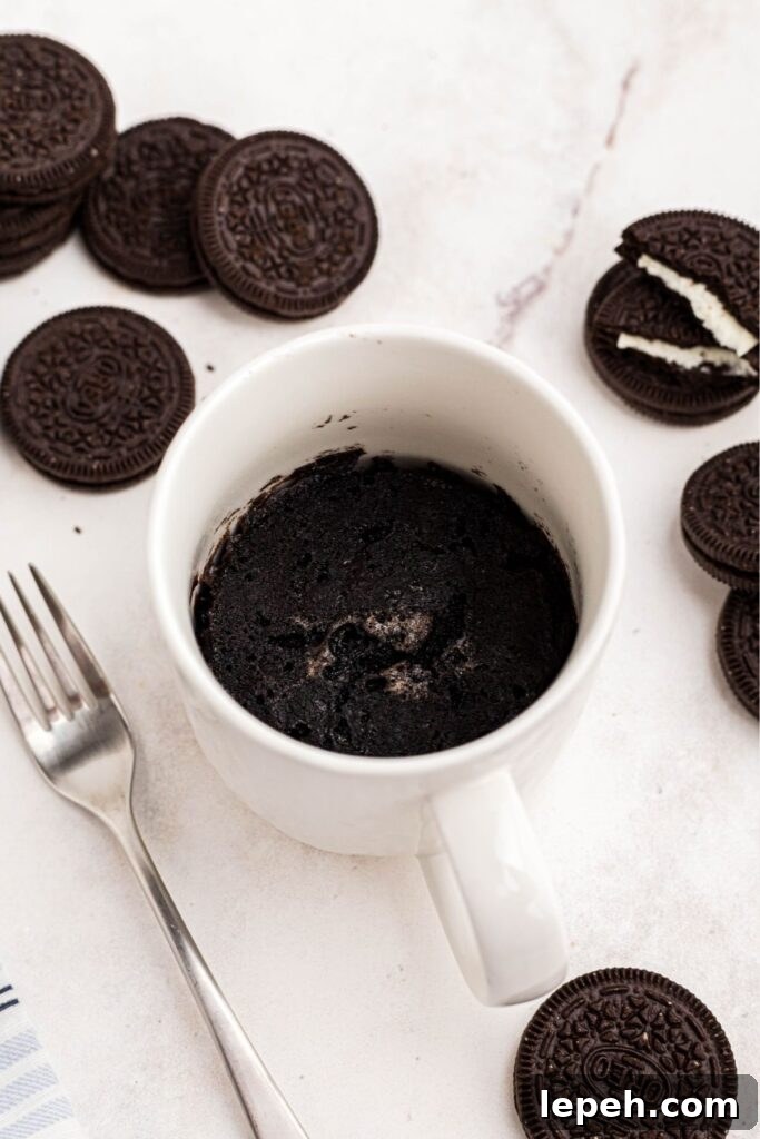 Chocolate cake cooked in a coffee mug, on a marble table with Oreo cookies scattered around the mug. 