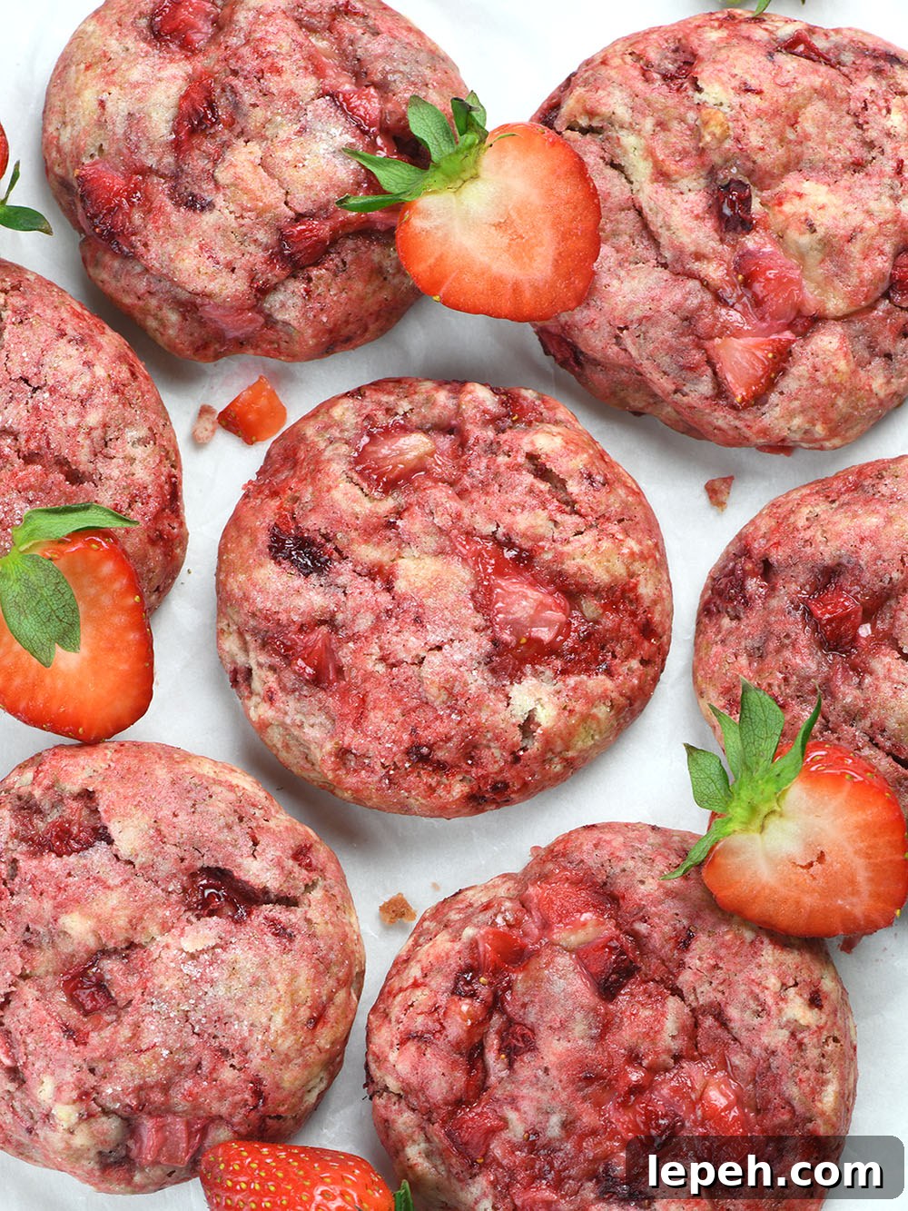 A top-down view of freshly baked strawberry cheesecake cookies with a vibrant pink hue, studded with juicy strawberry pieces, and lightly dusted with sugar. Fresh strawberry slices are scattered around.