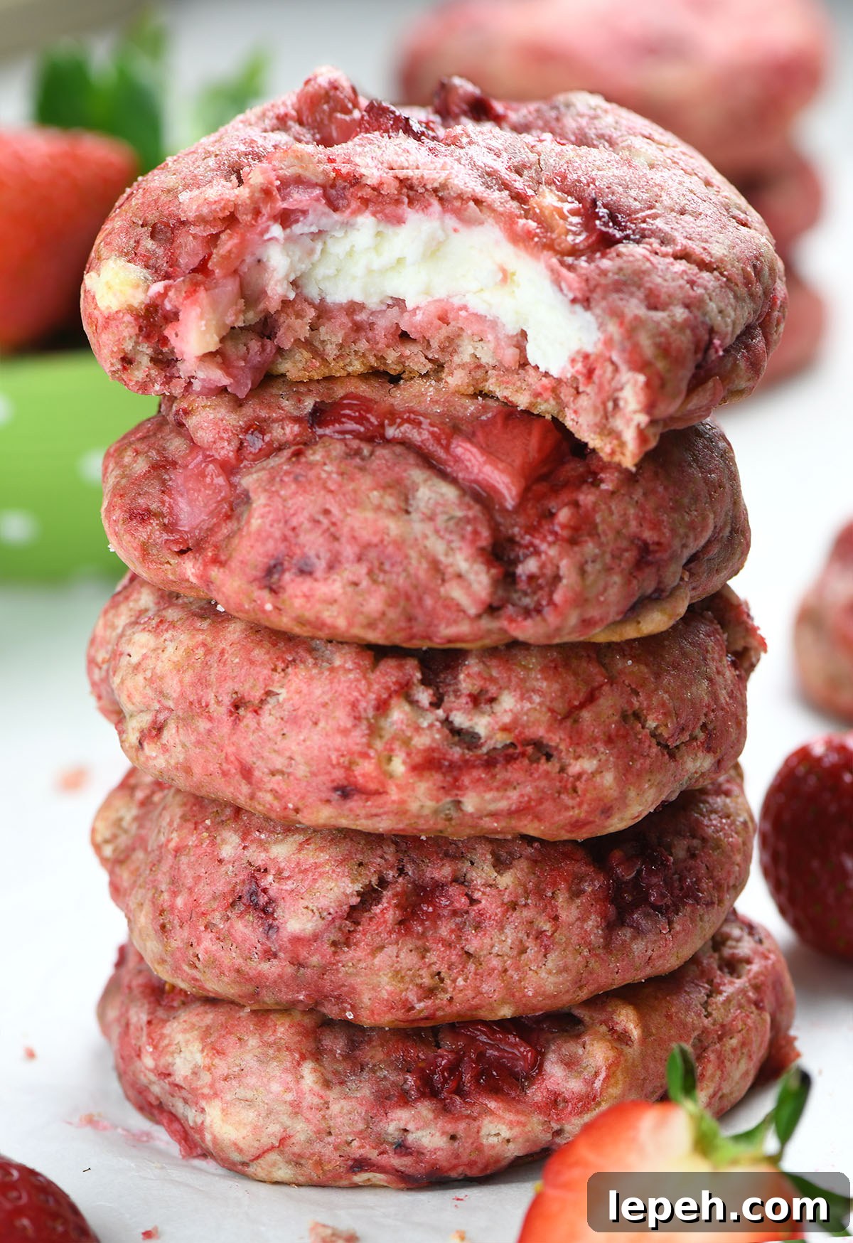A tall stack of strawberry cheesecake cookies with a bite taken out of the top one, revealing a creamy cheesecake filling. The cookies are vibrant pink with chunks of fresh strawberries, surrounded by whole and sliced strawberries.