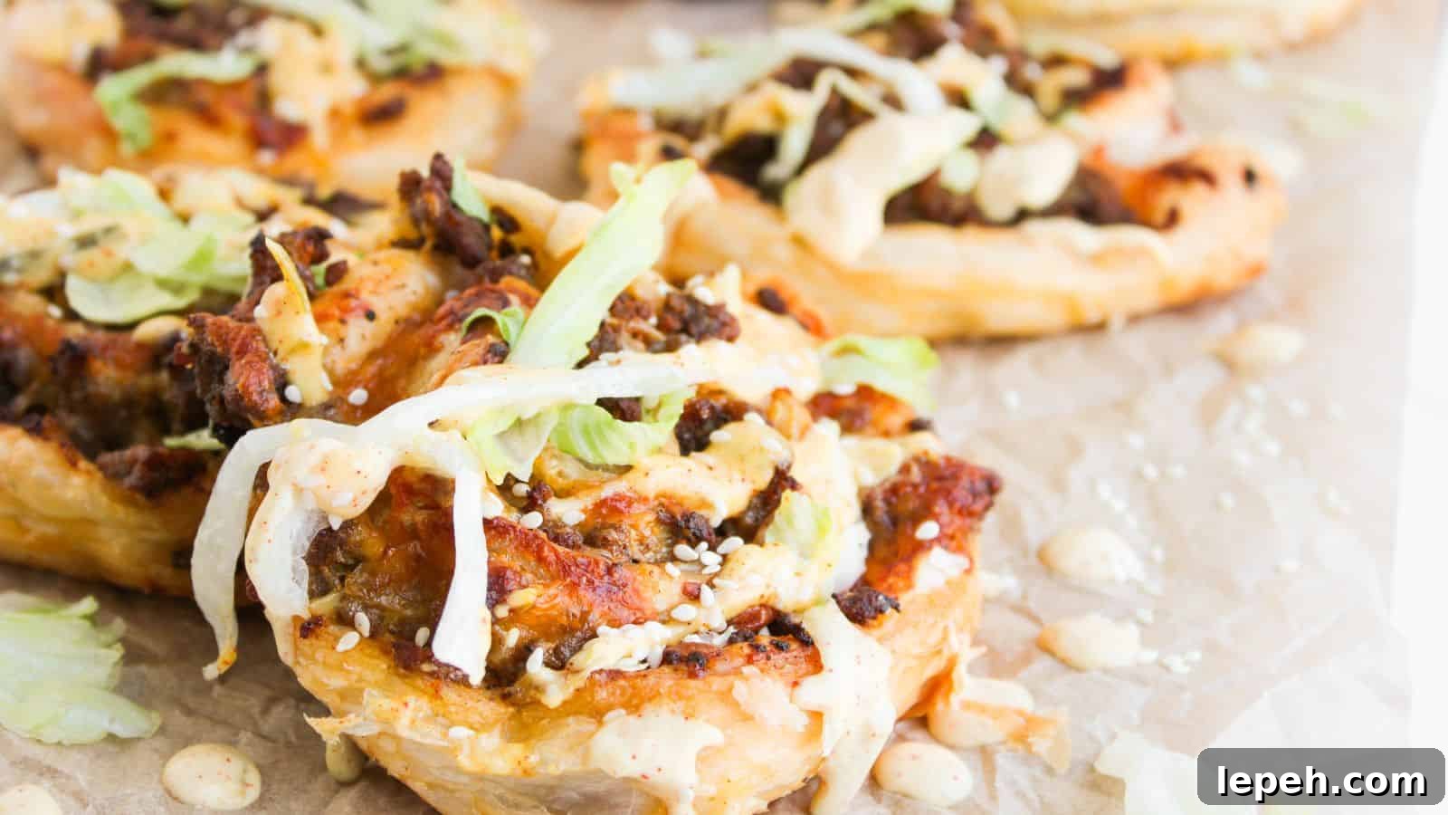Close-up of puff pastry rounds topped with ground meat, shredded lettuce, sesame seeds, and a creamy sauce, arranged on parchment paper.
