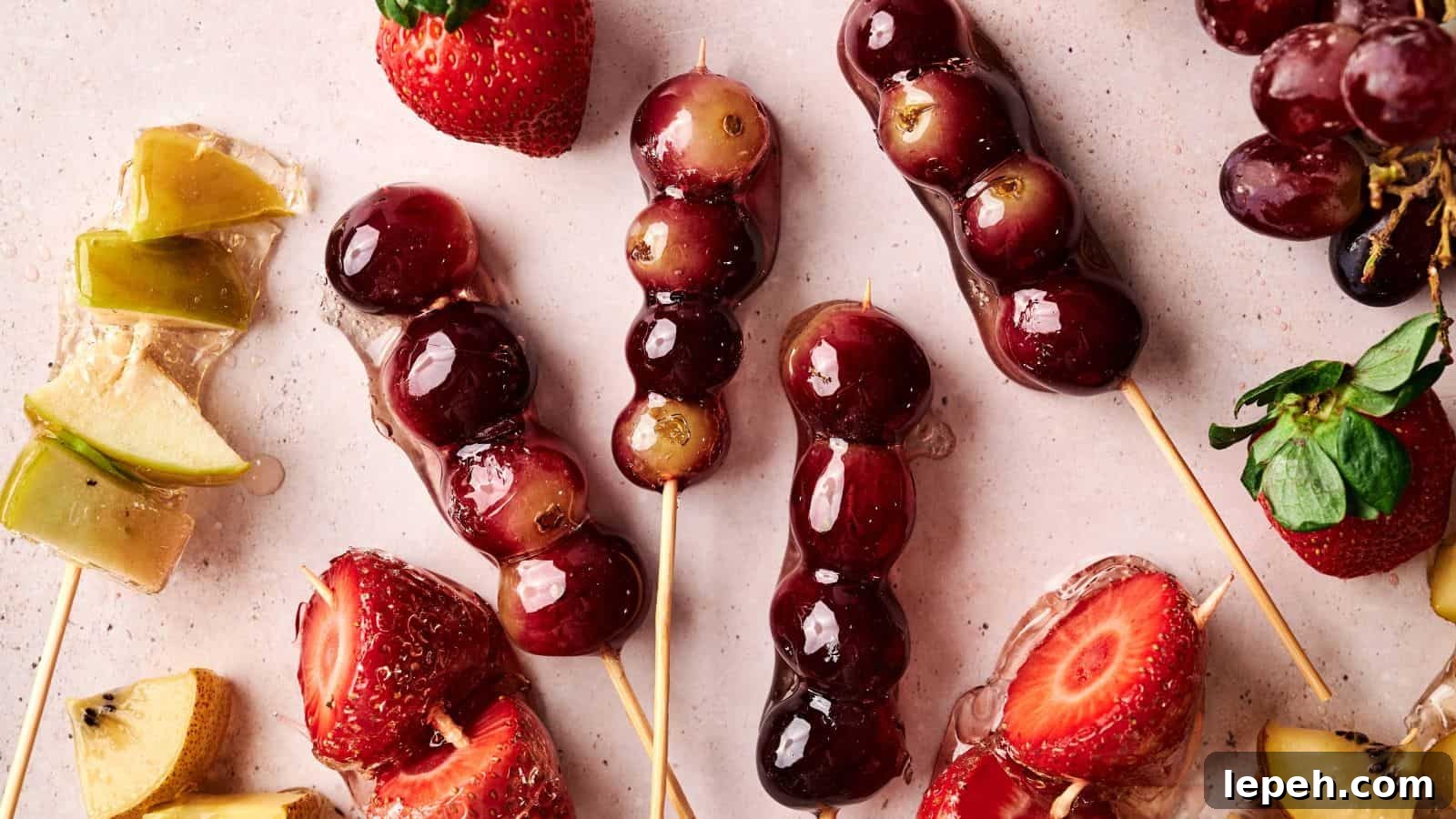 Skewers of grapes and strawberries coated in a shiny glaze are arranged on a light surface with loose fruit pieces nearby.