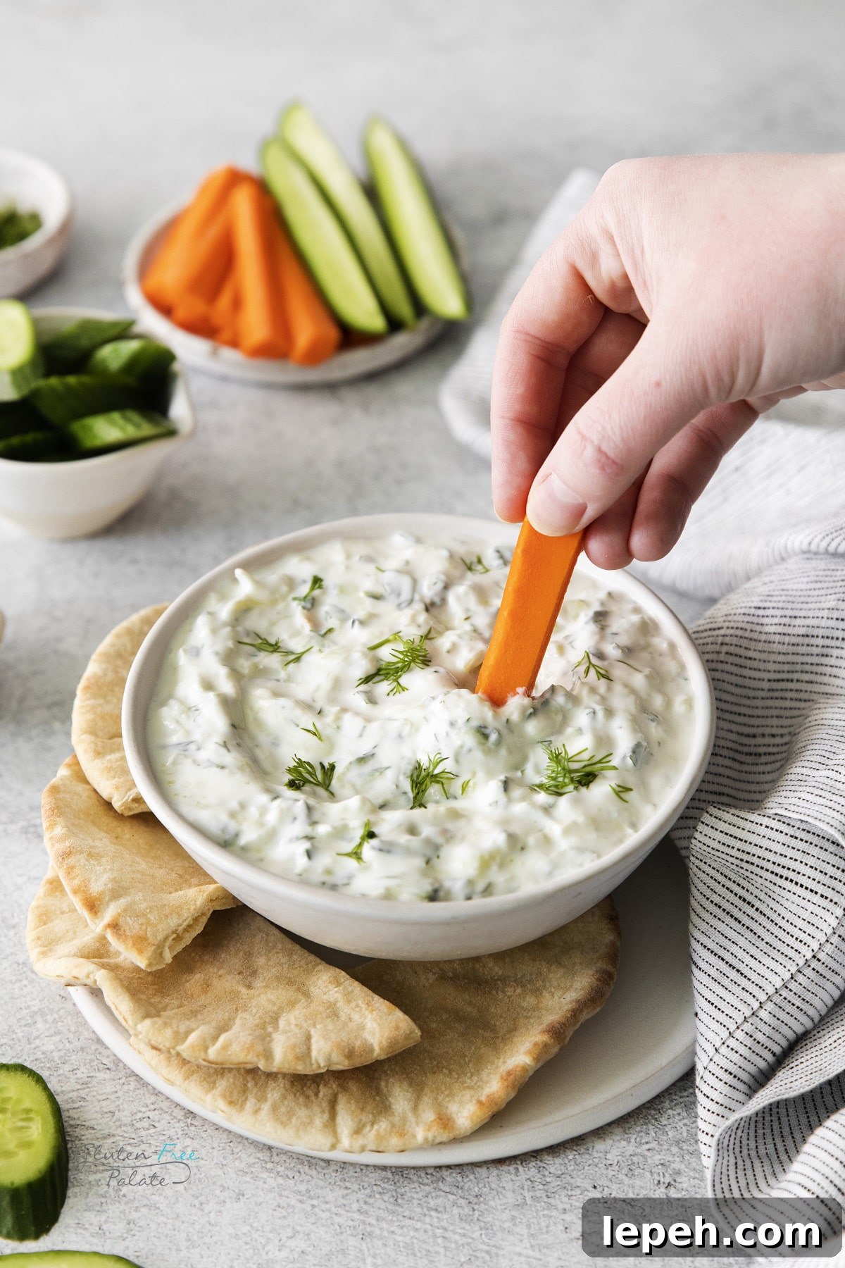 a white bowl of homemade tzatziki on a plate of pita bread. A hand is dipping a carrot stick into the dip