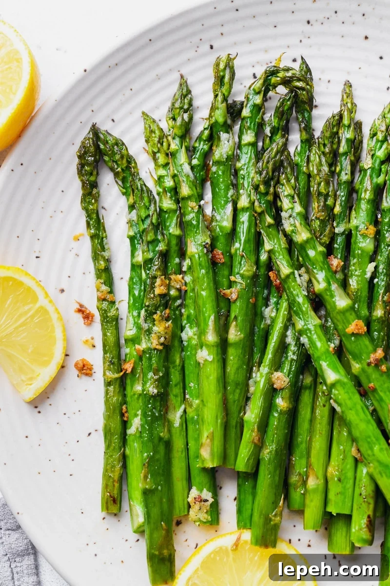 Overhead image of tender and caramelized bright green lemon garlic asparagus spears. On a large white speckled plate, next to a few slices of lemon. 