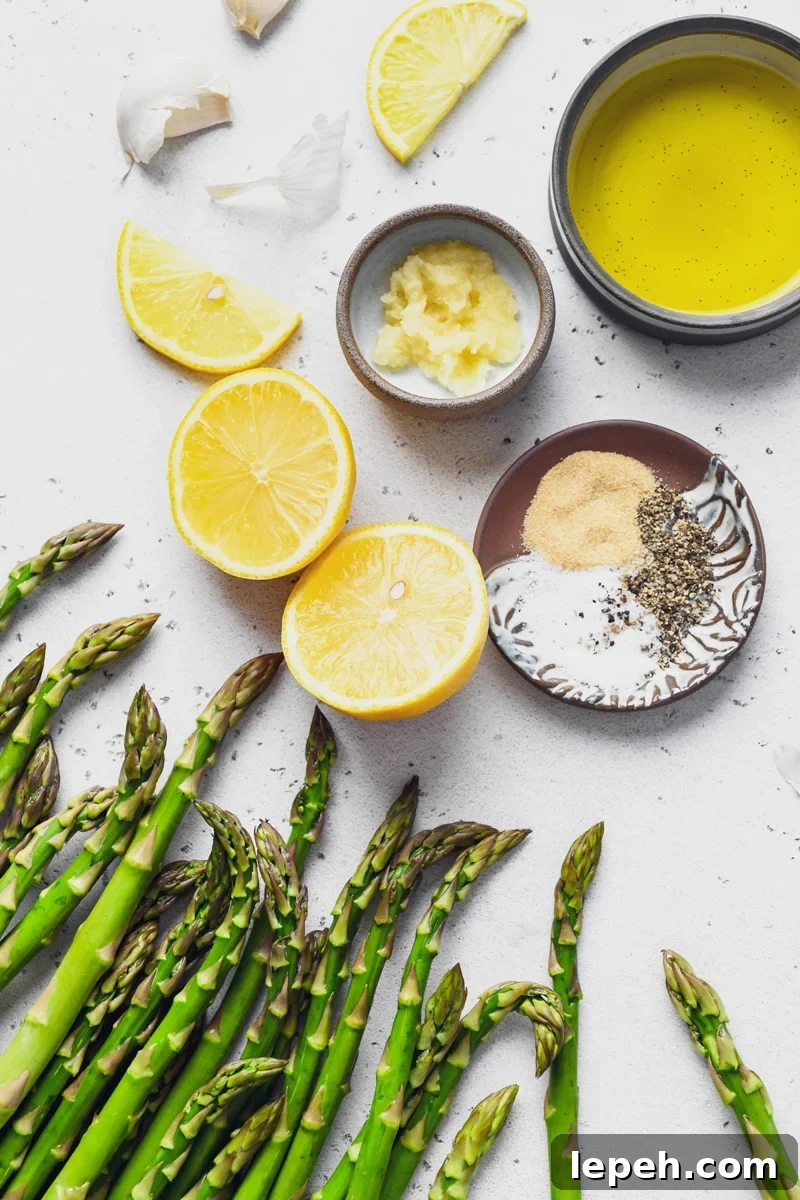 An overhead shot of scattered ingredients needed to make simple lemon garlic roasted asparagus. Asparagus spears, cut lemon, a small dish with grated garlic, another small dish with garlic powder, salt and pepper, and a bowl of olive oil. On a light colored speckled background. 