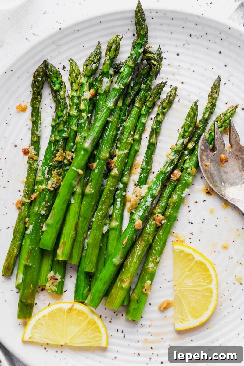 Overhead image of tender and caramelized bright green lemon garlic asparagus spears. On a large white speckled plate with a serving removed, next to a few slices of lemon. 