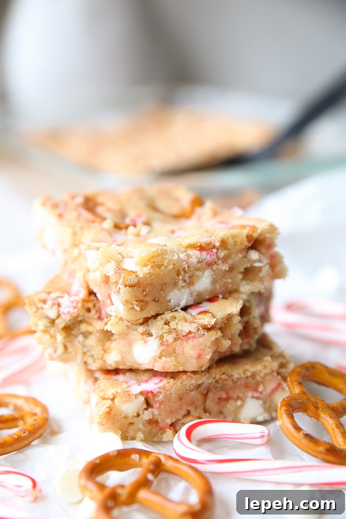 Three peppermint white chocolate pretzel cookie bars stacked on each other with small candy canes and pretzel twists surrounding them. Faded in the background is a glass 9x13 baking dish of the cookie bars and a black serving spatula.