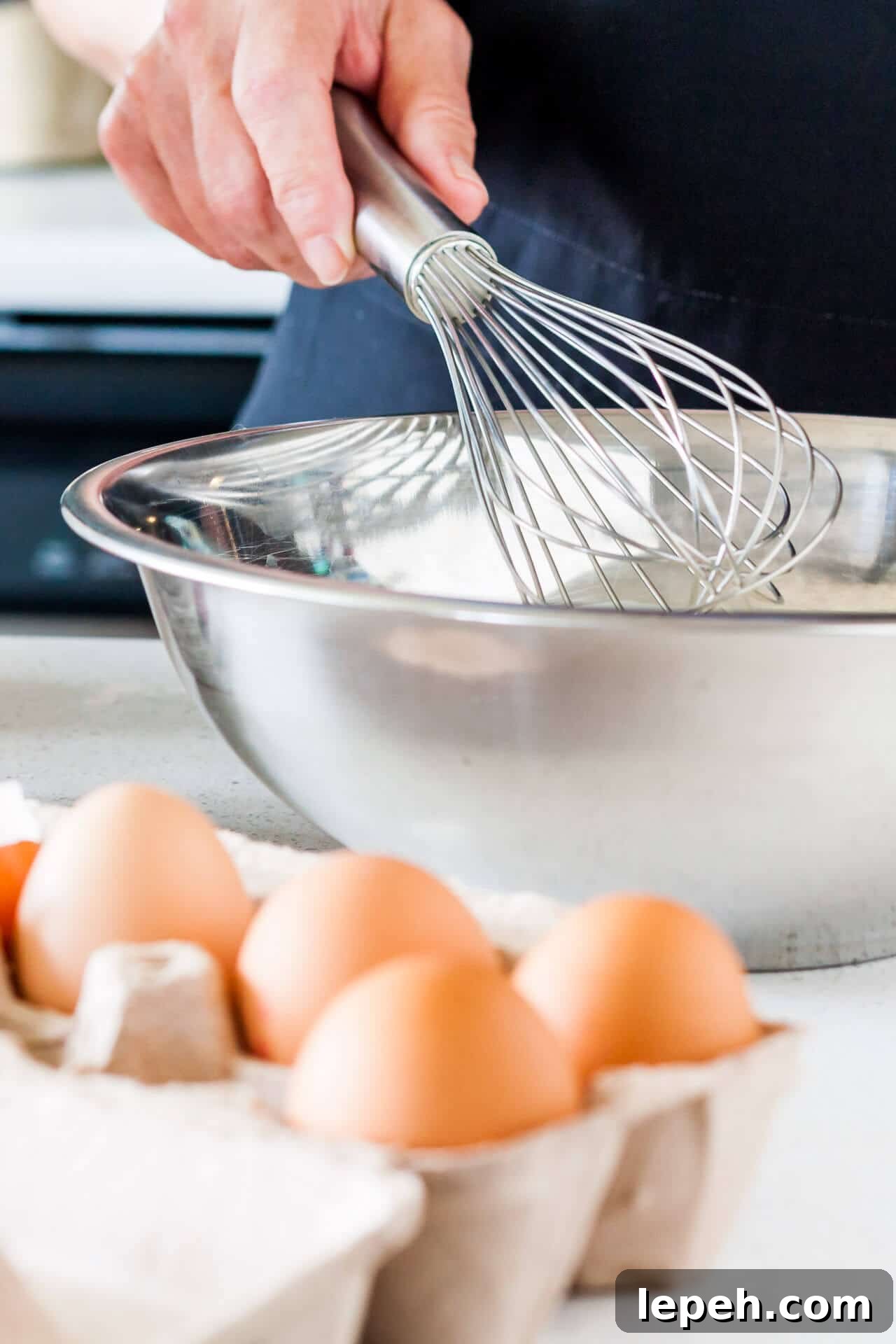 Whisking eggs by hand with a stainless steel whisk in a stainless steel bowl with a carton of brown eggs displayed next to the bowl