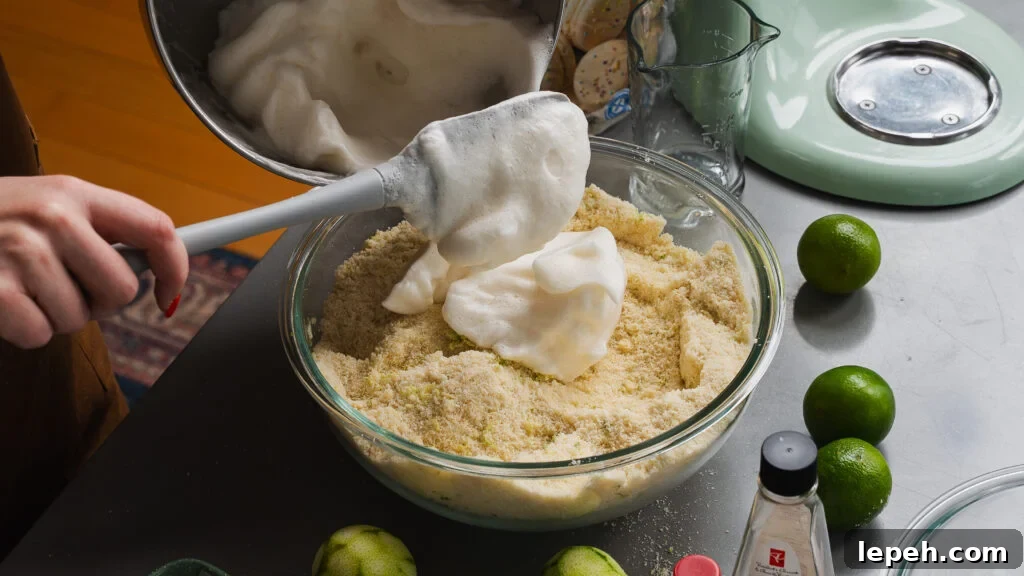 Adding egg whites with a spatula to an amaretti cookie base in a clear bowl.