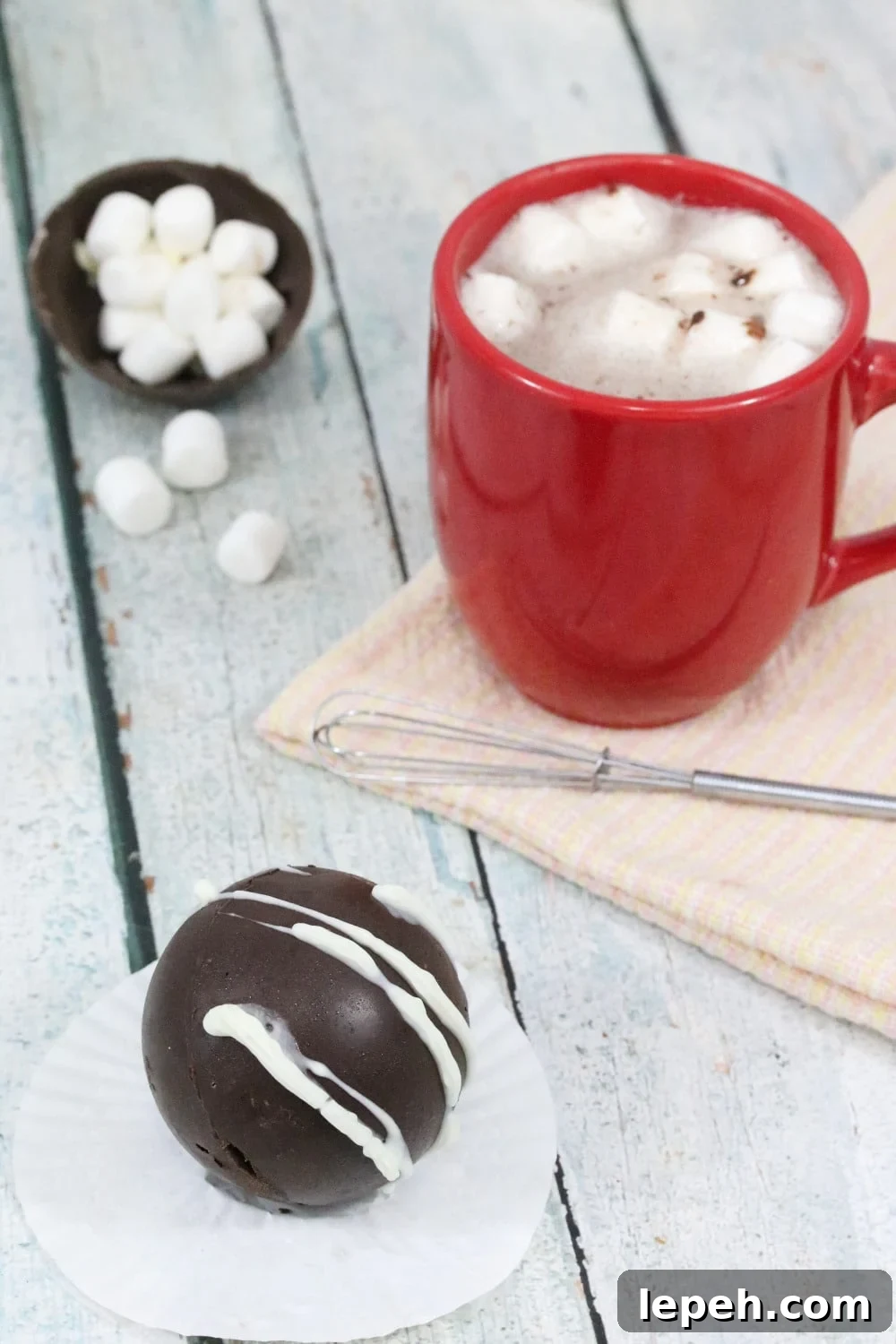 hot chocolate bomb on a white cupcake liner with more bombs in a white bowl behind it and a red mug of hot chocolate with a whisk and marshmallows