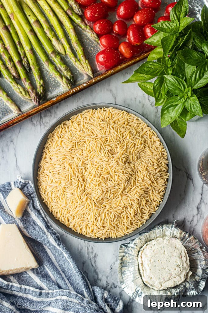 Raw orzo, raw asparagus and tomatoes in a pan, parmesan cheese on blue tea towel, basil leaves on marble board, boursin cheese unwrapped on countertop.