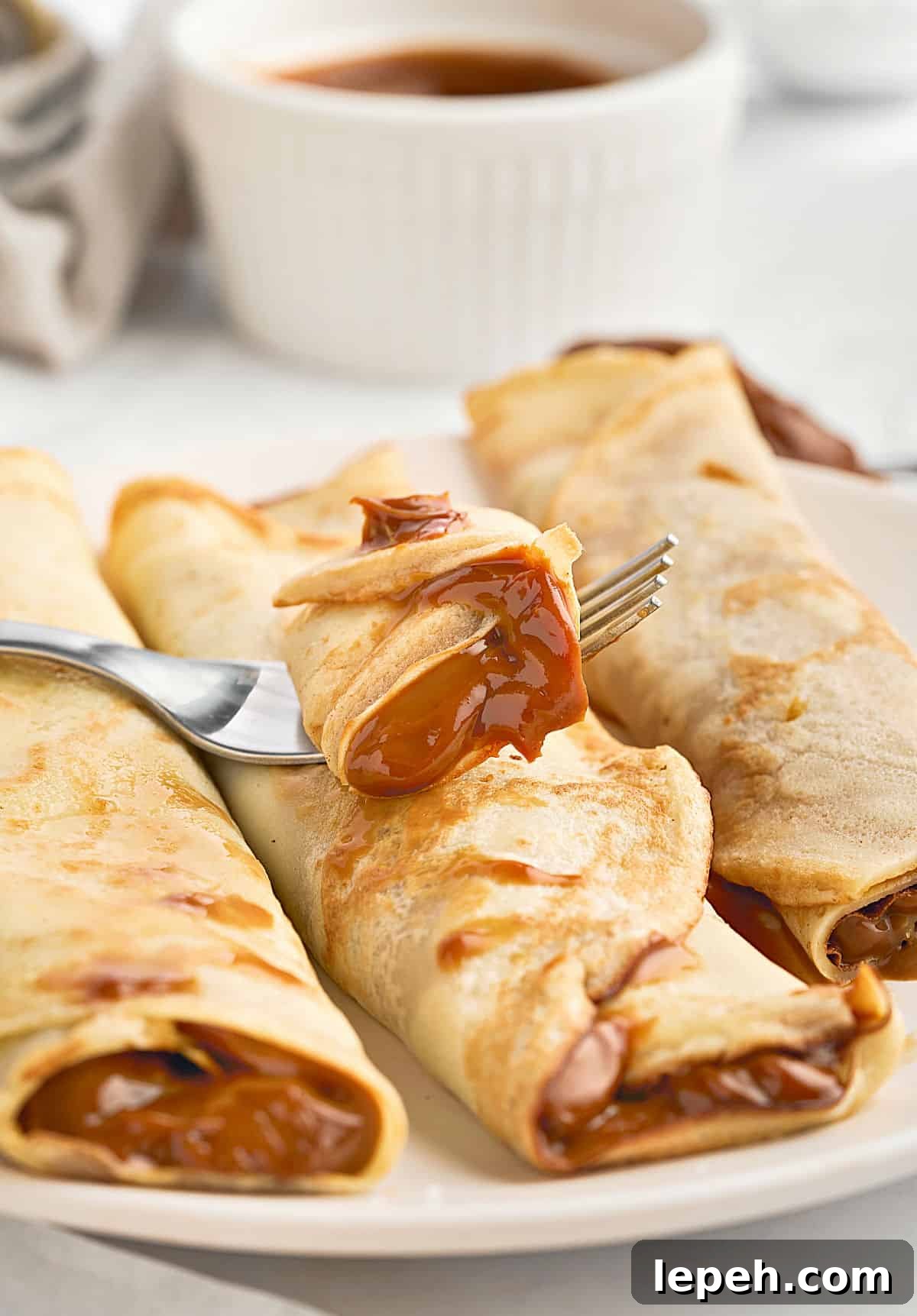 Forked bite of dulce de leche crepe on more rolled pancakes. White plate and background.