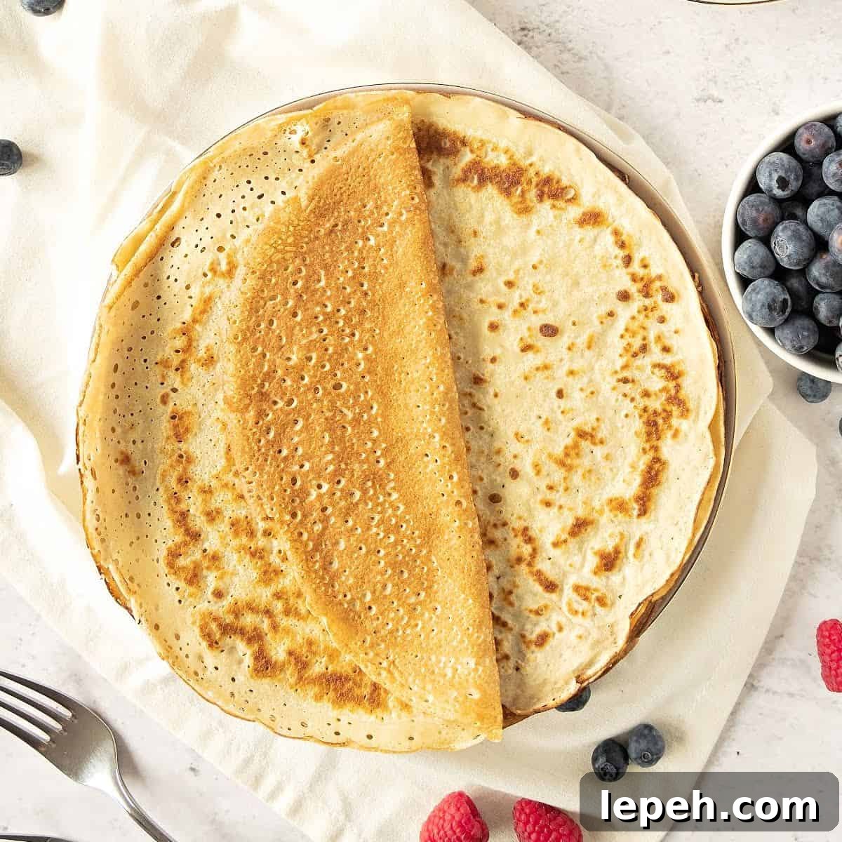 Whole thin pancakes on a plate. White towel and surface. Whole berries around. Top view.