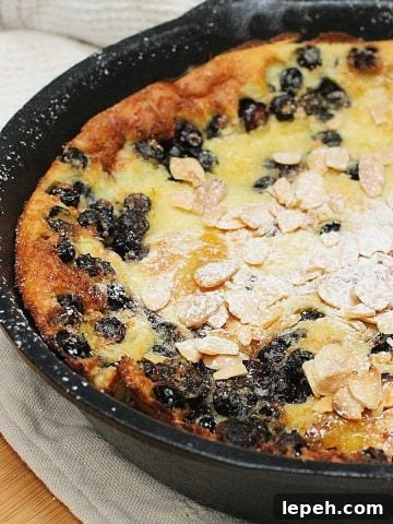 Cast iron skillet with partial view of blueberry white chocolate pancake. Beige cloth and wooden board below.