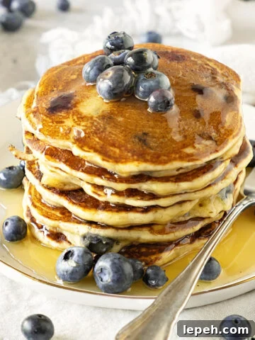 Several stacked ricotta blueberry pancakes with syrup. White plate. Silver fork.