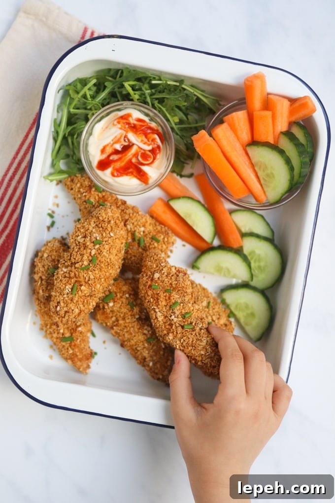 Oat Crusted Chicken Goujons in a white enamel serving dish with a side serving of chopped carrots and cucumbers and a glass ramekin filled with a dipping sauce.