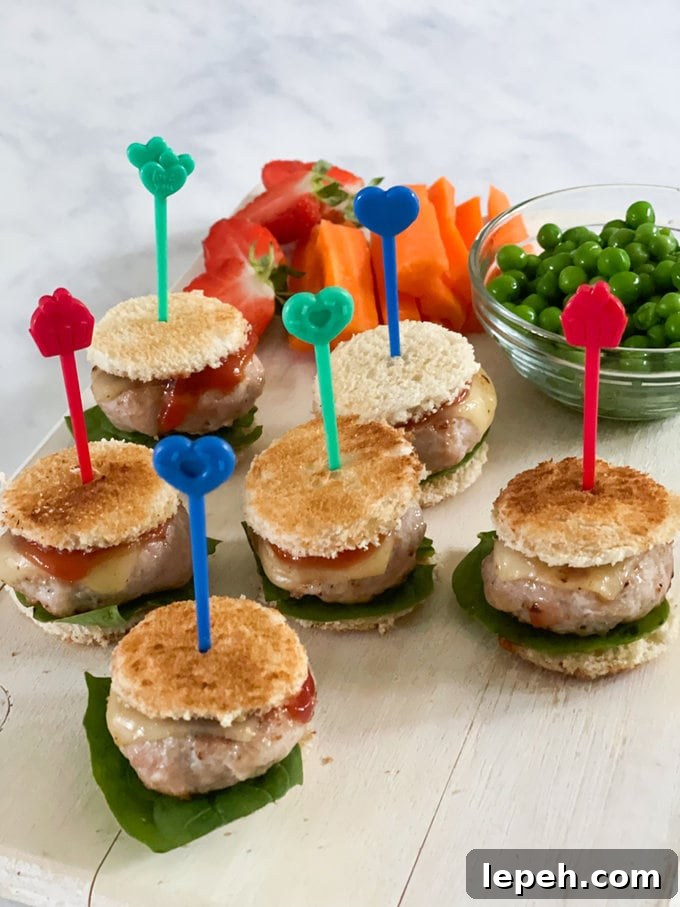 Mini Chicken Burgers served held together with Pick Sticks and served on a wooden chopping board with a side of peas, carrot batons and chopped fruit.