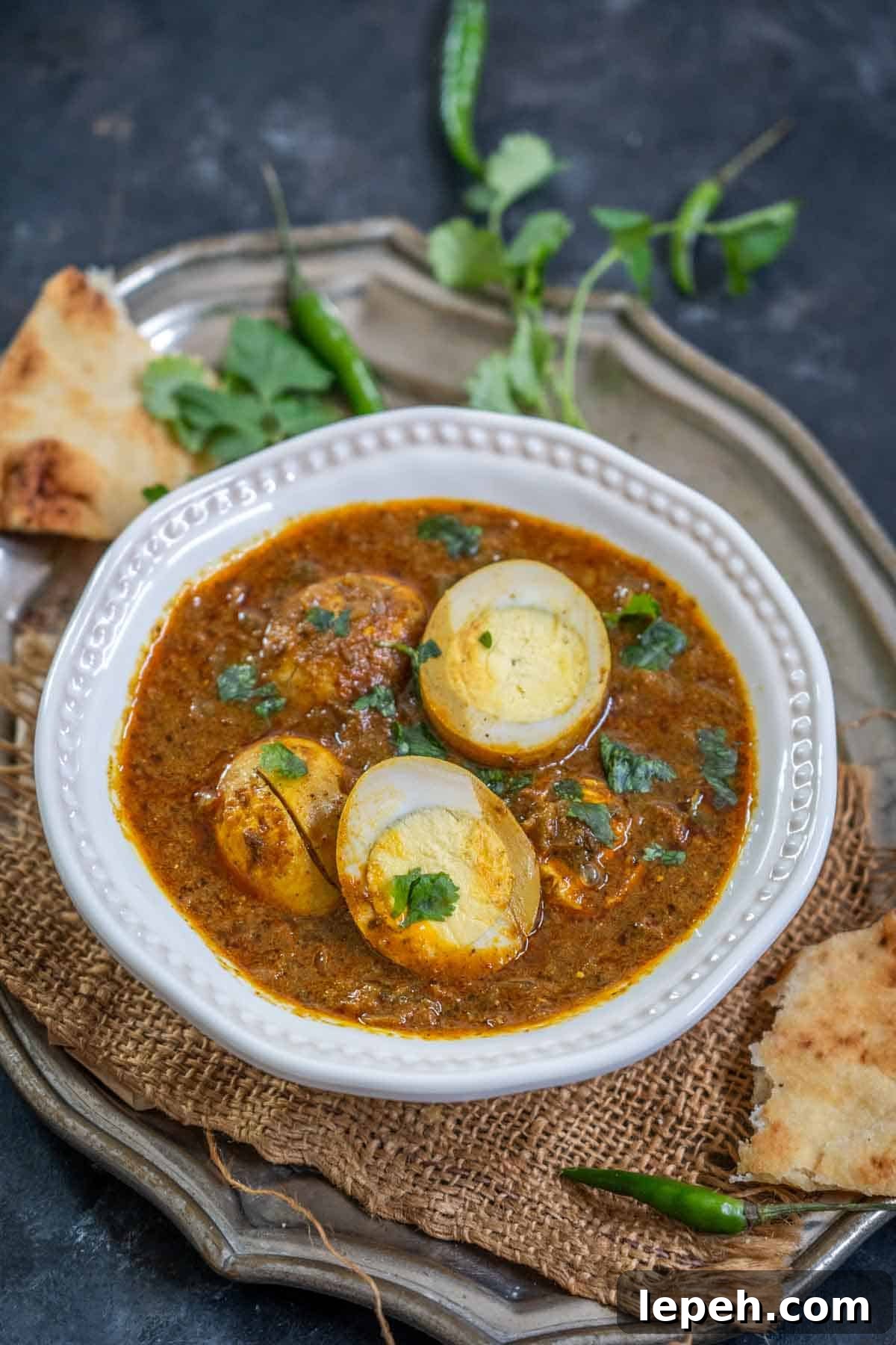 A white bowl of Dhaba Style Egg Curry garnished with cilantro, served with pieces of naan bread and green chilies on a rustic metal tray, set against a dark background.
