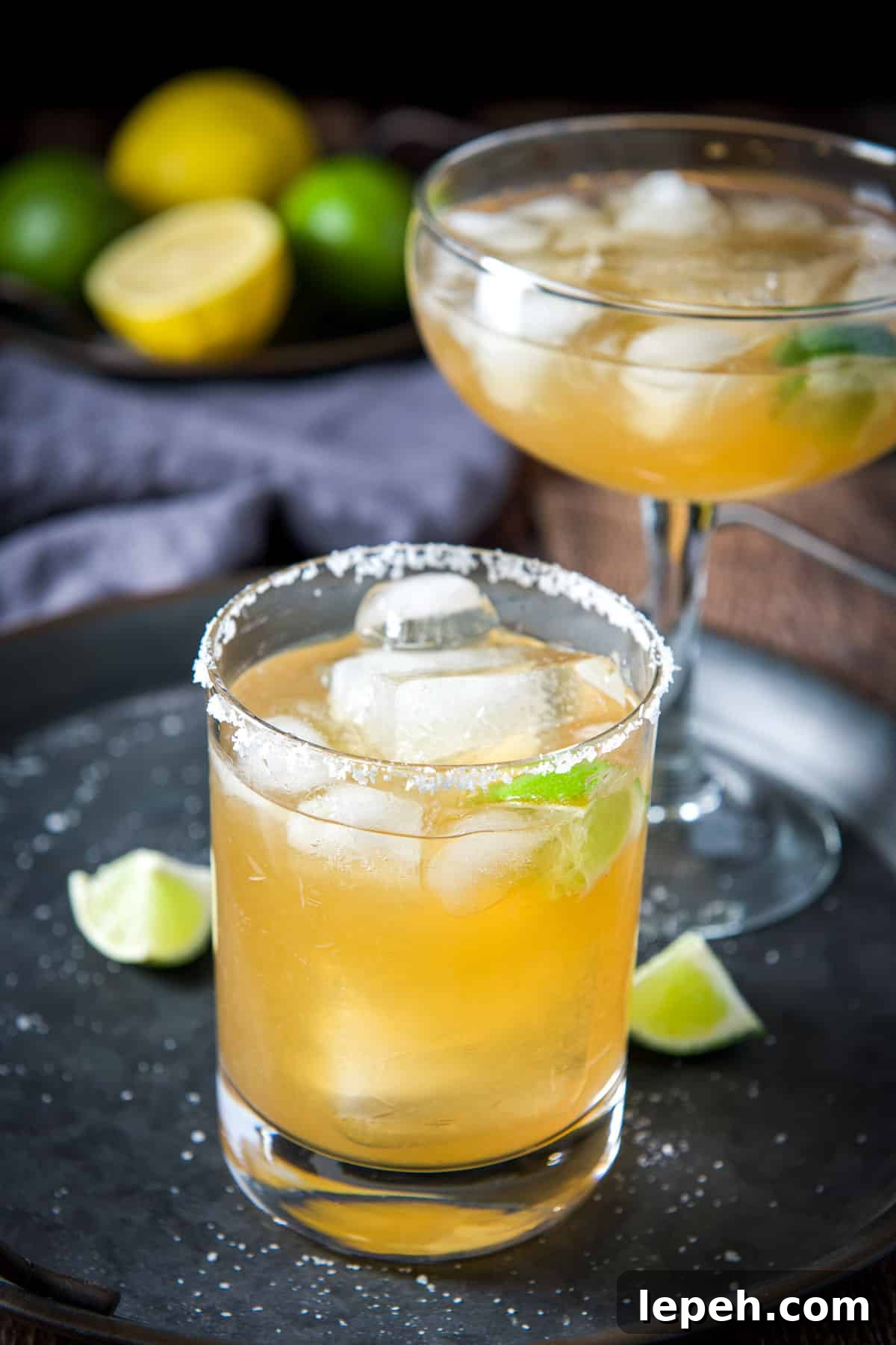 A salted glass in front of a bowl glass with margaritas in them with lemon and lime behind