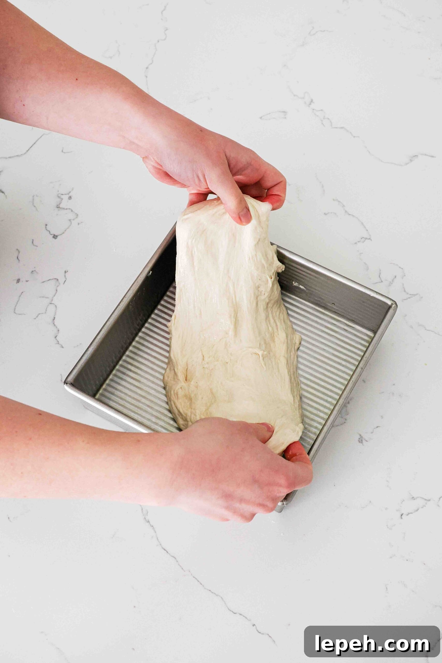 Two hands stretch dough over a square 8"/20 cm pan.