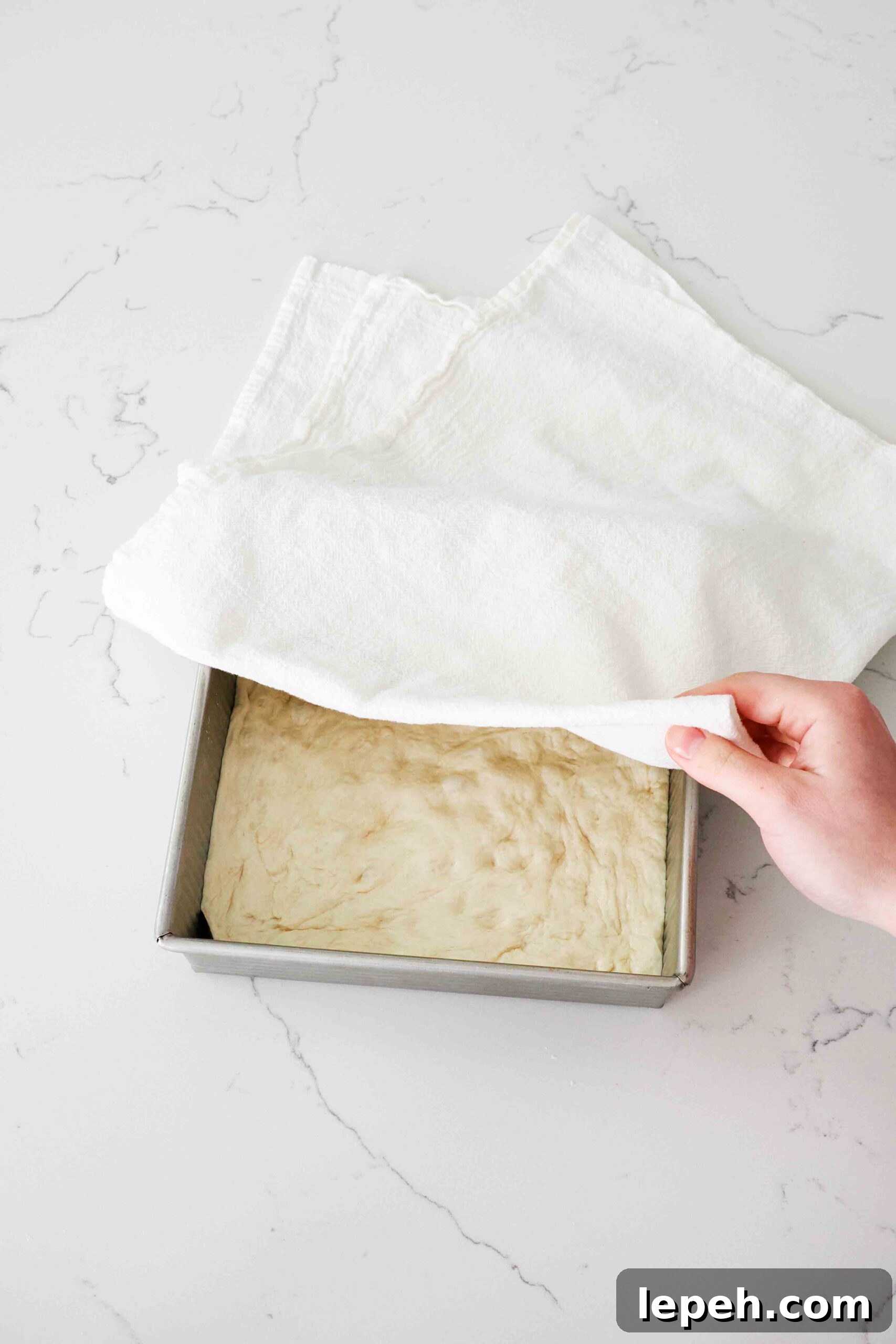 A hand pulls a kitchen towel over a square pan with focaccia dough in it.