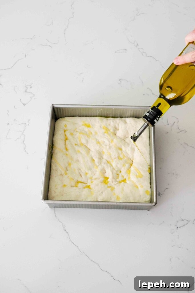 A bottle of olive oil drizzles oil over a small batch of focaccia dough.
