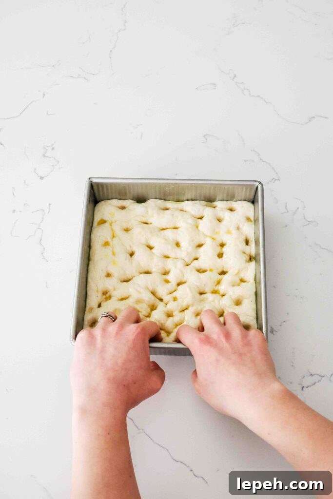 Two hands press their fingertips into proofed focaccia dough in a square pan.