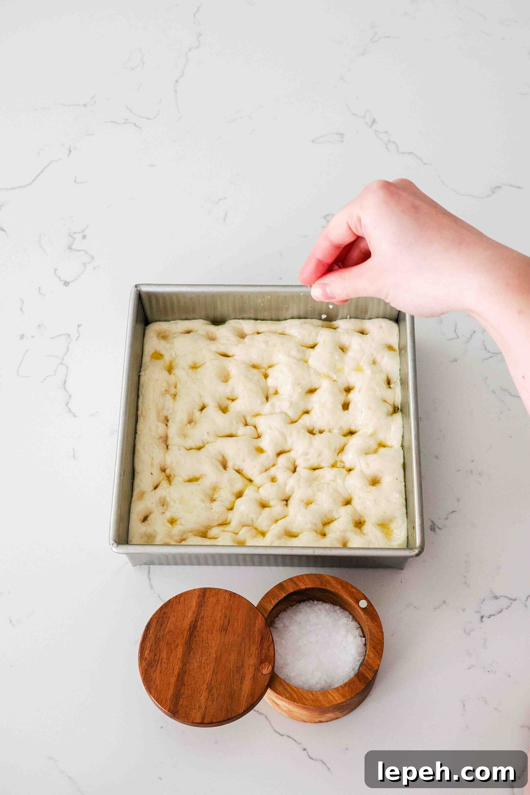 A hand sprinkles flaky salt over an unbaked loaf of focaccia bread.