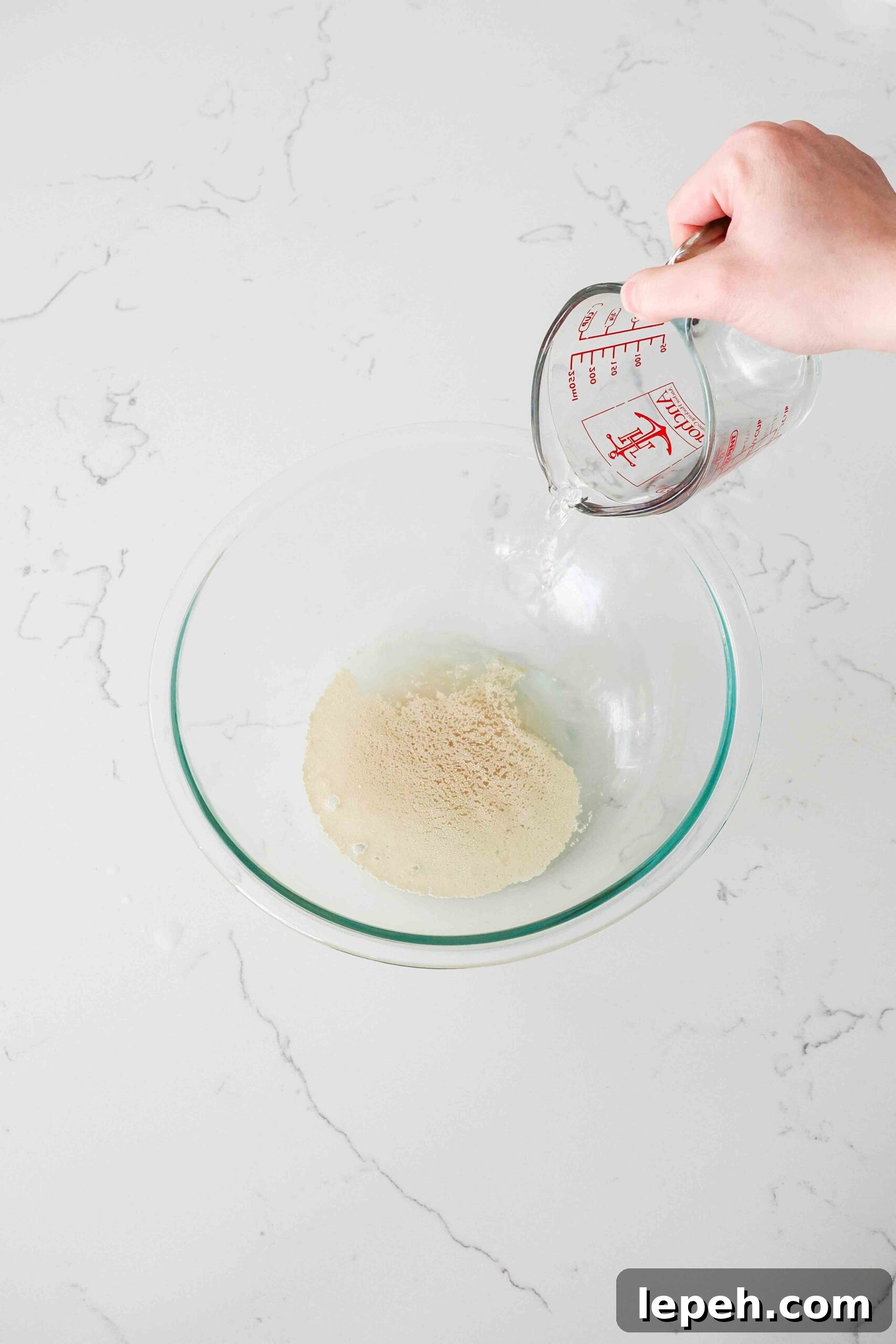 A hand pours warm water from a measuring cup over yeast in a glass mixing bowl.