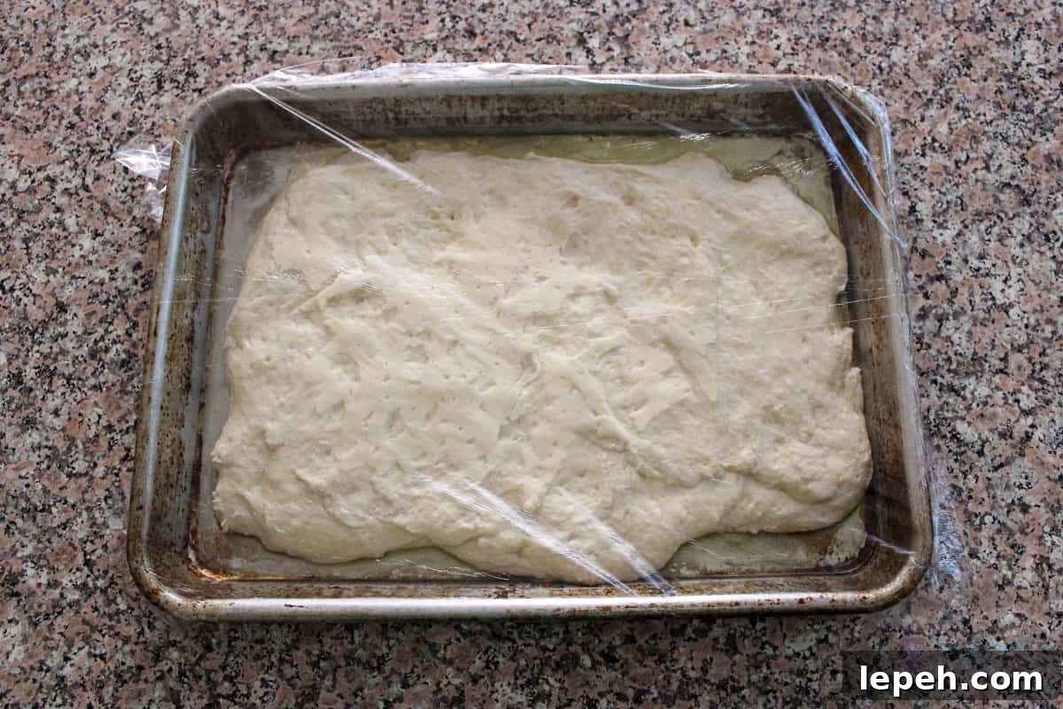 Pizza dough on quarter sheet pan with plastic wrap over it.