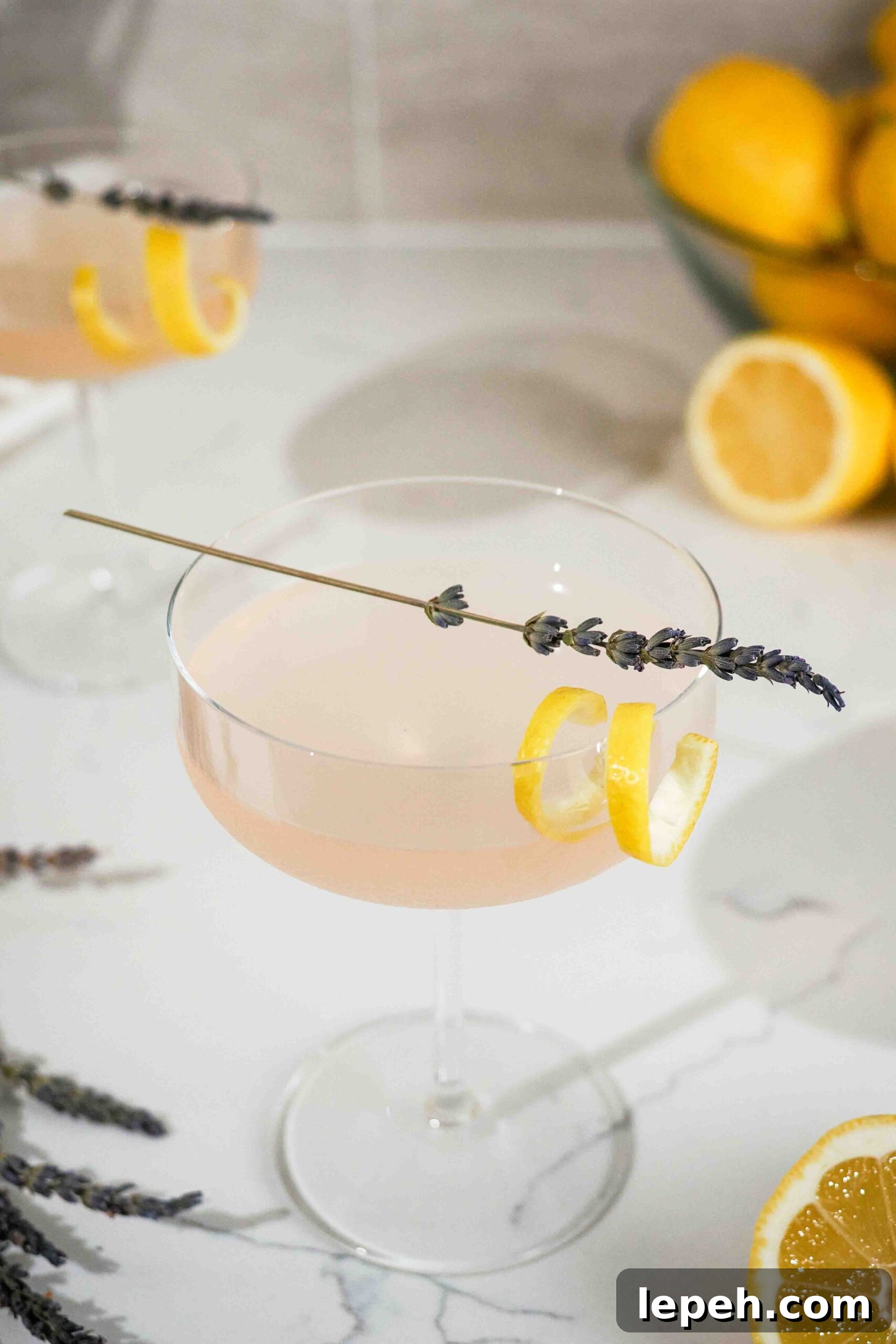 A lavender lemon drop martini in a coupe glass on a counter with lemons.